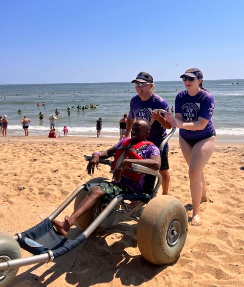 A young man with paraplegia been pushed out of the ocean by two people while sitting in a beach wheelchair with oversized back wheels.