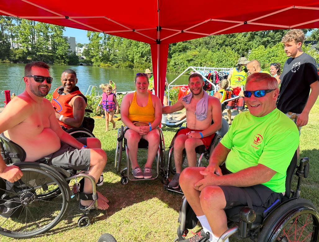 Five people all in wheelchairs sitting under a red tent in their beachwear on a very sunny day, with the lake behind them.
