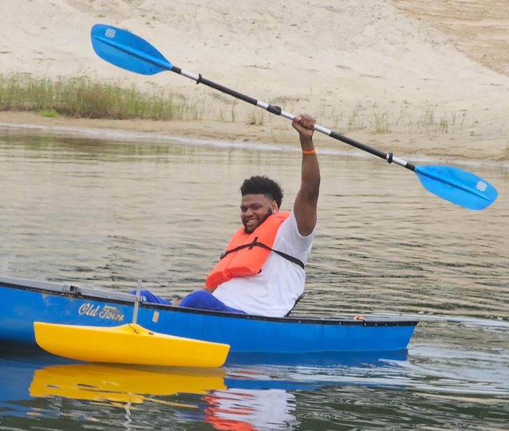 A young man with paraplegia smiling in a canoe, & holding a double-headed paddle high above his head.