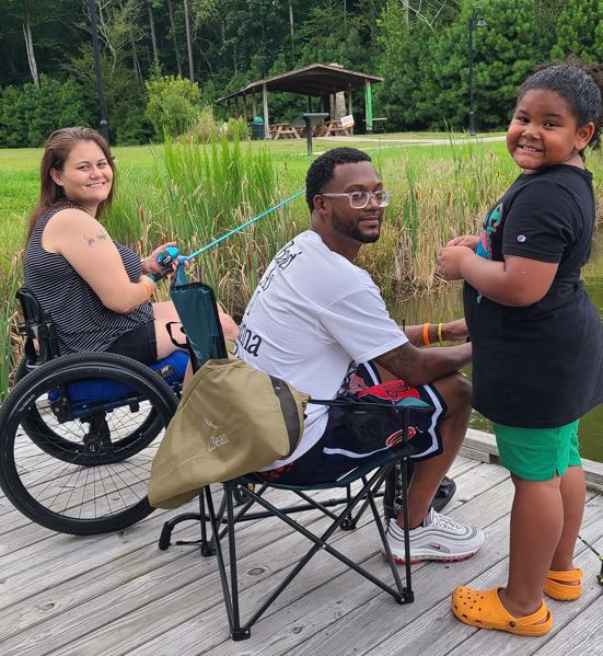 A female paraplegic sitting on a dock fishing while sitting by her husband, who is sitting in a folding chair, while their daughter stands next to them holding bait.