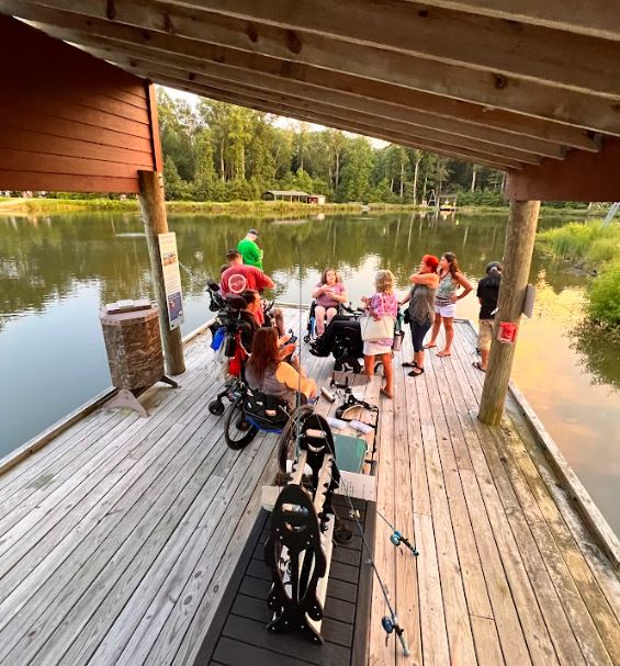 A group of wheelchair users and people who can stand all together at the end of a wood covered pier fishing and talking with one another.