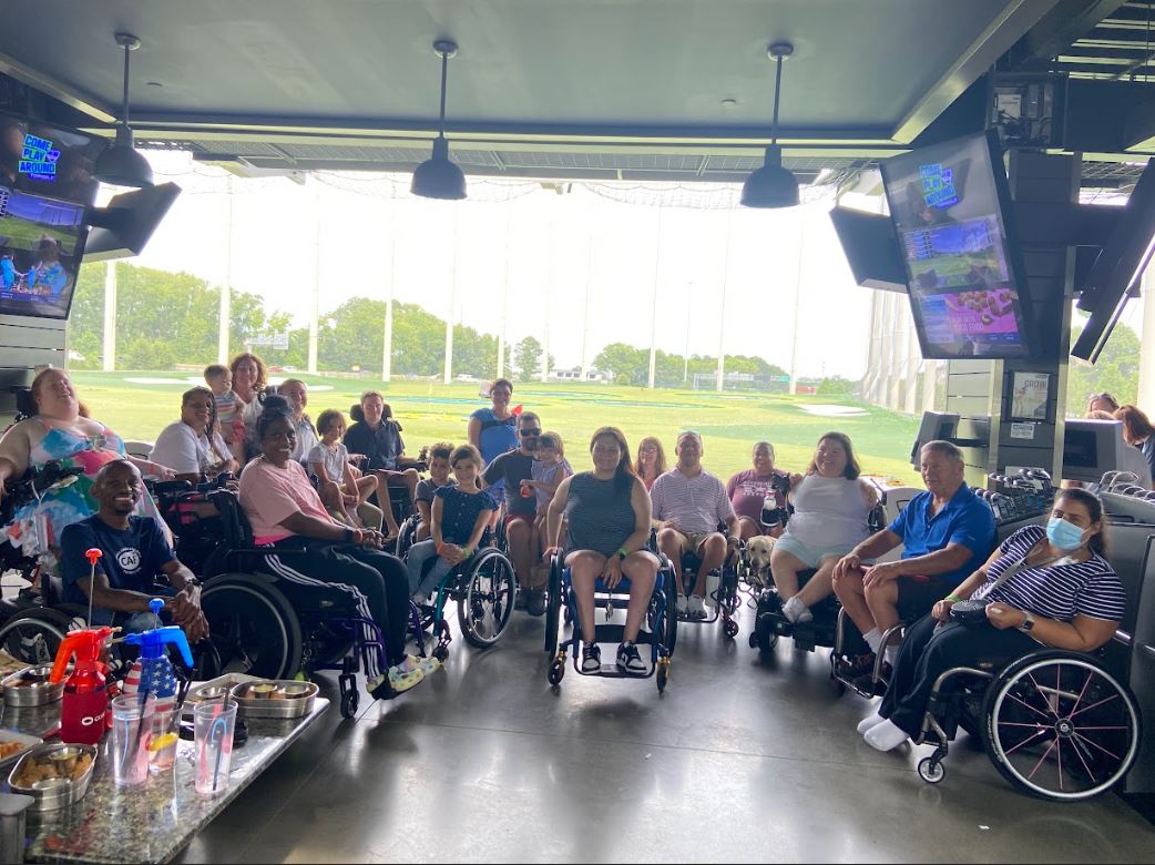 A group of around 25 people, mostly wheelchair users, sitting together at Topgolf in a rented pod, with the driving range behind them.