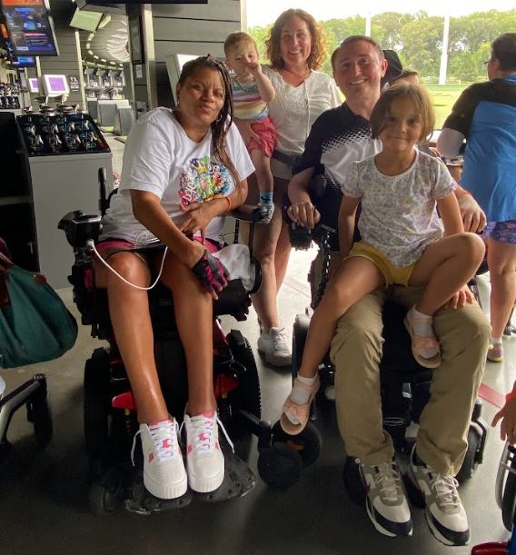 Josh sitting in his wheelchair with his daughter on his lap, with his partner standing behind him holding their younger son.  Another woman in a wheelchair sits next to Josh on his left side. 