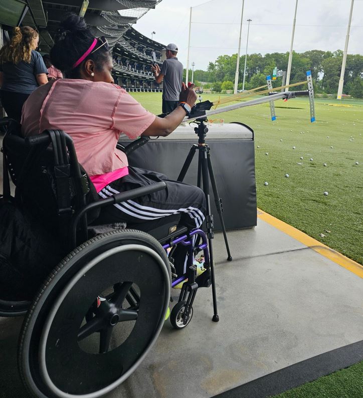 Woman sitting in wheelchair pulling on slingshot loaded with a golf ball.
