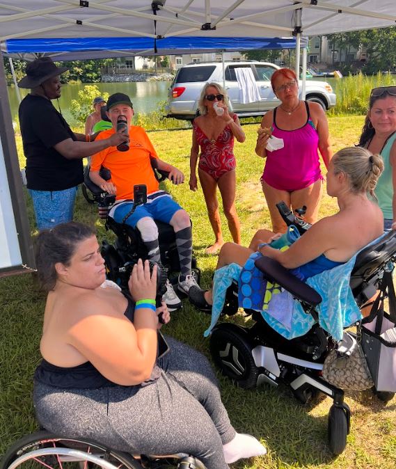 Three wheelchair users and four able bodied people hanging out under a tent drinking water and relaxing.