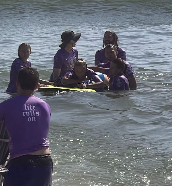 A woman with quadriplegia lying on her stomach on a surfboard while floating in shallow water, while five able bodied people stand around her making sure the surfboard stays stable.