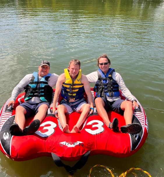A blond hair man with paraplegia sitting on a bright red flotation device in the water, with two able bodied people sitting on either side of him.  Everyone is wearing a life jacket.